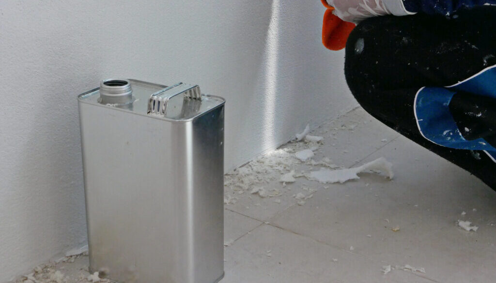 A gallon of thinner placed next to old concrete wall, while a painter is removing sticky rough glue and tape remain on the wall, as a preparation before starting the paint the house