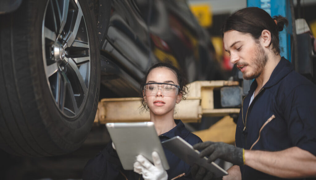 Young male and female car mechanic wearing uniform with safety eye glasses and hand gloves working on digital tablet while diagnosing problem in car